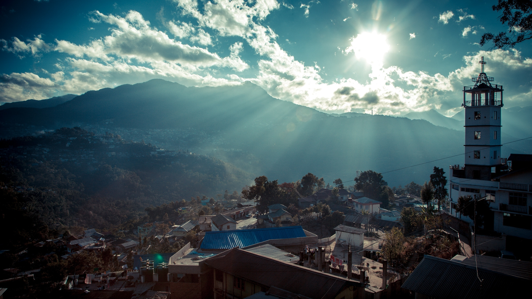 Sun breaking through clouds over a hillside town with a white church tower and cross