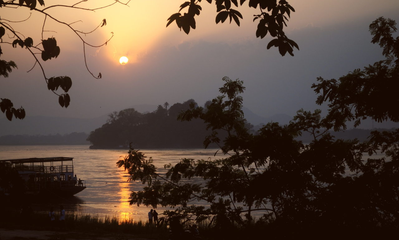 Sun setting over a wide river, with a moored passenger ferry and people on the bank