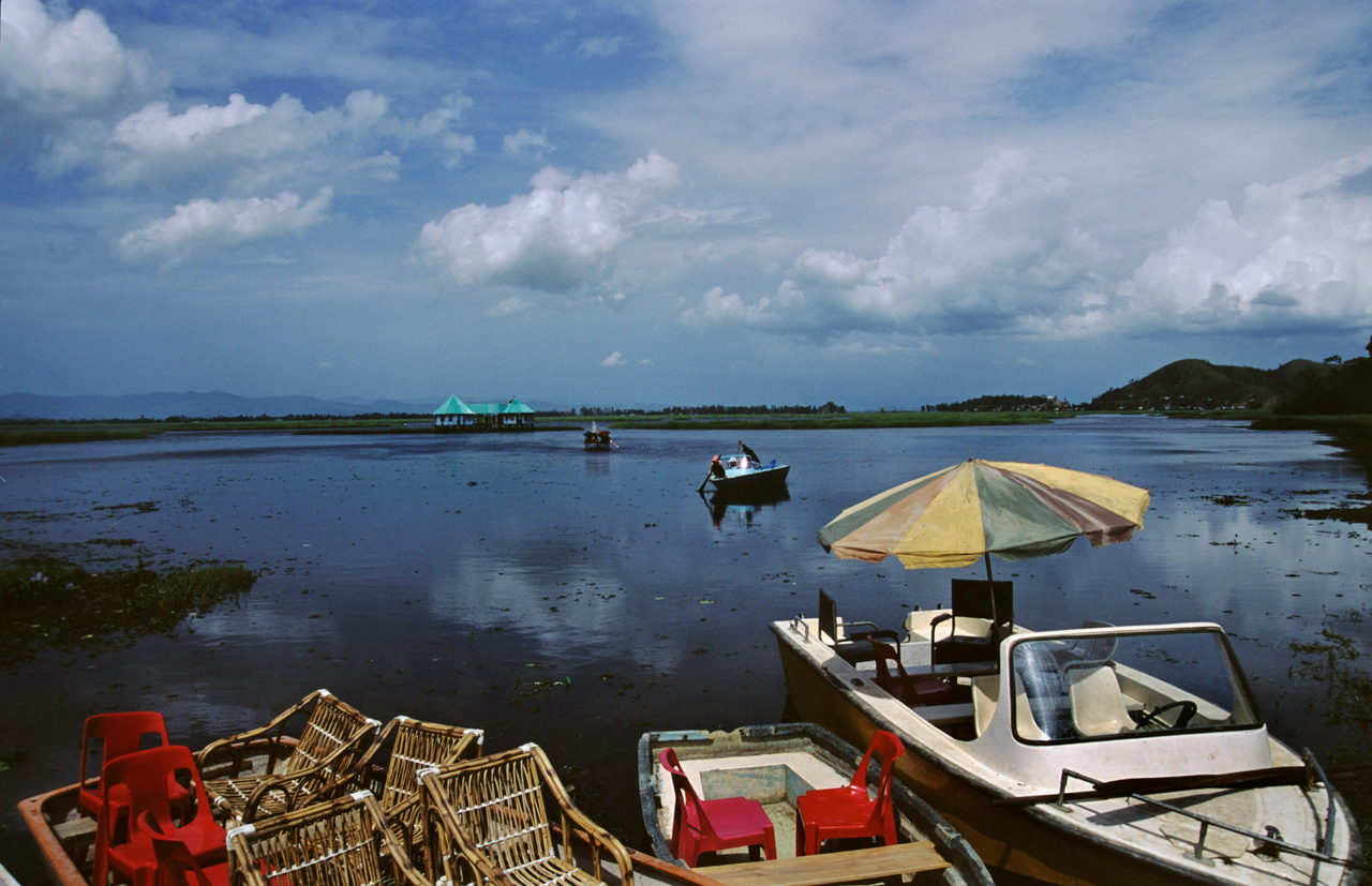 Moored boats with red plastic chairs and a faded umbrella on a wide lake
