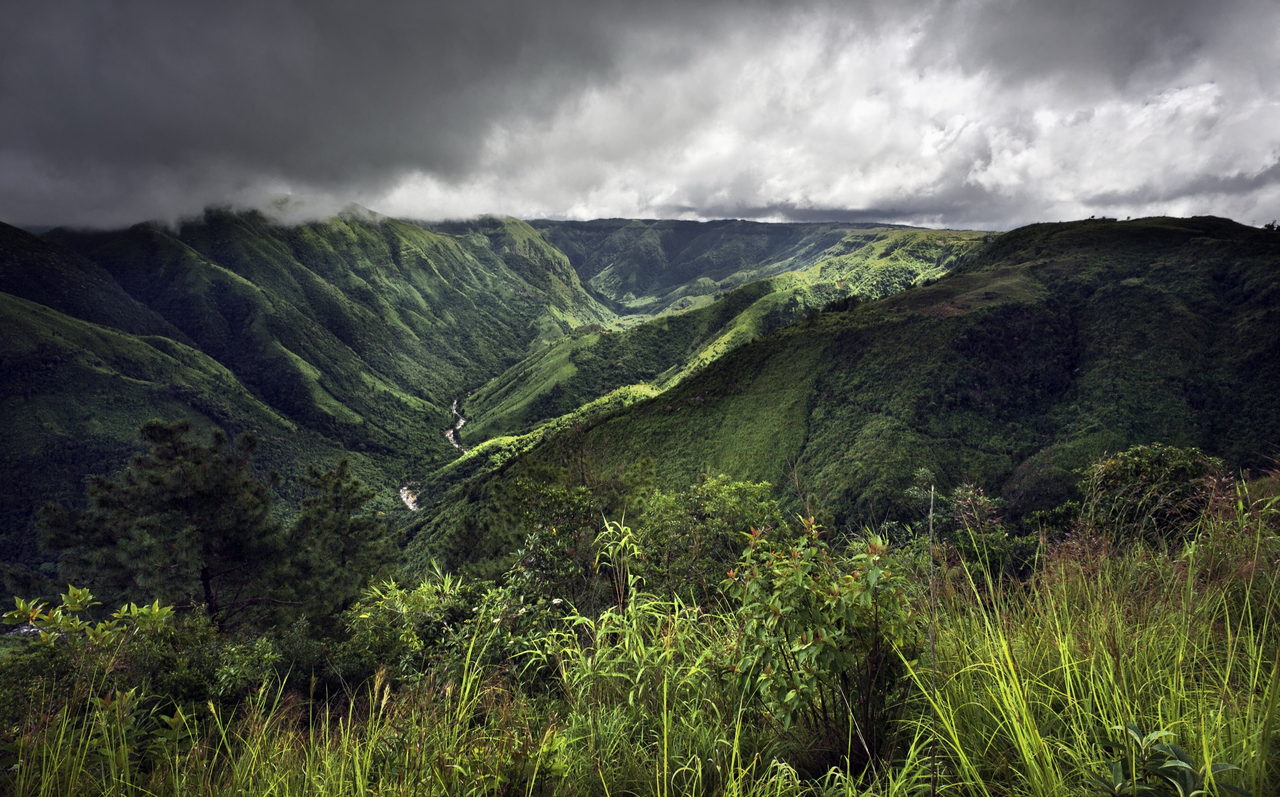 Steep green valley under heavy storm clouds, with a stream running through the gorge below